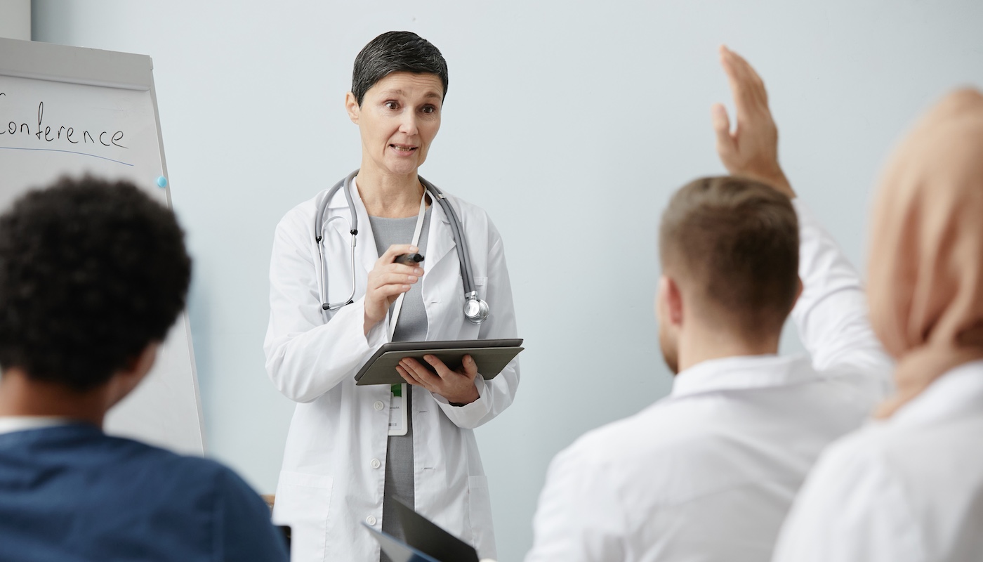 Teacher Wearing Stethoscope and Holding Clipboard Answering Questions from Seated Students